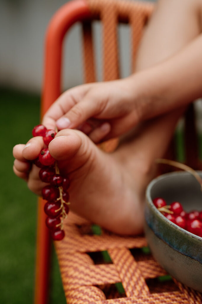 Hand hält Johannisbeeren zwischen Zehen auf einem orangenen Stuhl