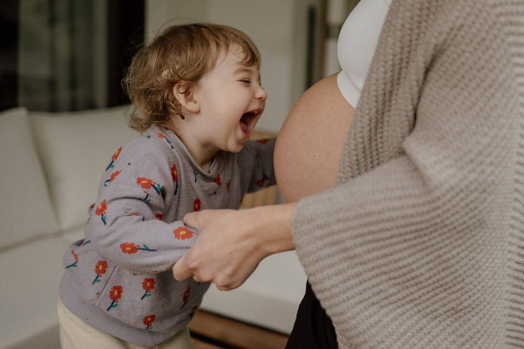 Kleinkind hält Hände von Mama und lacht Babybauch an