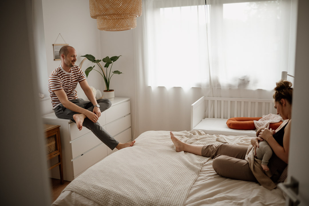 Baby wird im Schlafzimmer gestillt während Papa auf Kommode sitzt und zuschaut