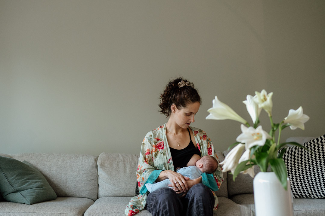 Mama sitzt auf Couch und stillt Baby neben Blumen